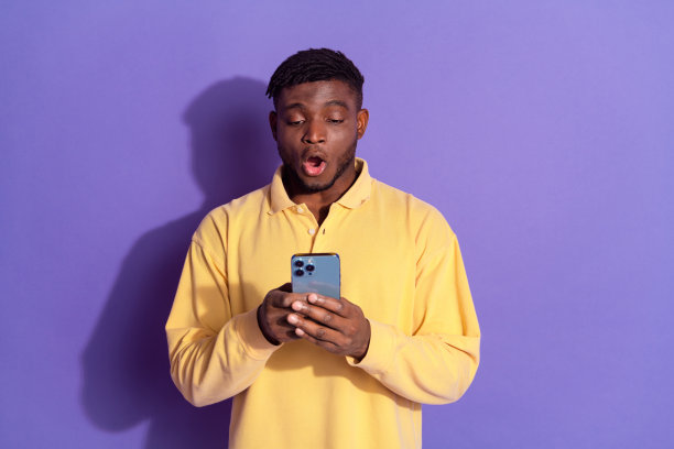 漂亮的年轻男性的照片肖像震惊地拿着装置，穿着时髦的黄色衣服，孤立在紫色的背景上图片下载