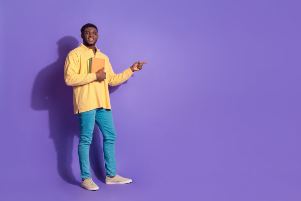 迷人的年轻人手持书籍点空的全身照片穿着时髦的黄色衣服孤立在紫色的背景图片下载