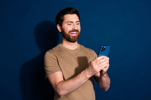 照片中的年轻帅哥黑发模特穿着米色t恤用智能手机交流孤立的深蓝色背景图片下载