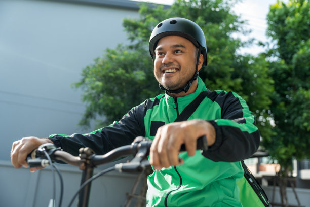 肖像骑手送餐员穿着绿色制服戴着头盔骑着自行车送餐给顾客。快乐快递员用绿色背包将货物运送给客户。图片下载