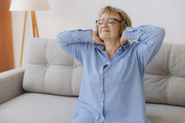 患病的老年成年妇女坐在沙发上摸脖子有肩颈疼痛。保健和医药理念图片下载