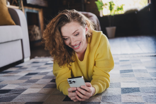 迷人的年轻女性躺在地板上的全尺寸照片，手持装置穿着黄色毛衣舒适的日光家庭室内客厅图片下载