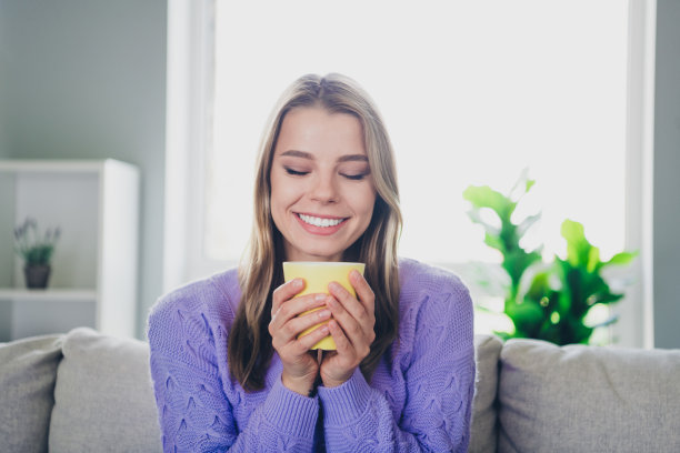 迷人的年轻女子的照片坐在沙发上喝咖啡杯闻香气穿着紫色的衣服舒适的日光家室内客厅图片下载