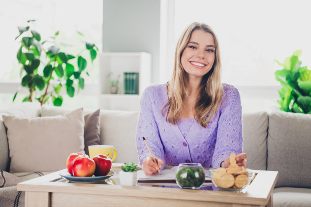 迷人的年轻女子的照片坐在沙发桌子写笔记本穿着紫色的衣服舒适的日光家室内客厅图片下载
