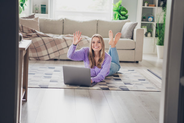 漂亮的年轻女子躺在地板上的全尺寸照片上网本挥手hi穿着紫色的衣服舒适的日光家室内客厅图片下载