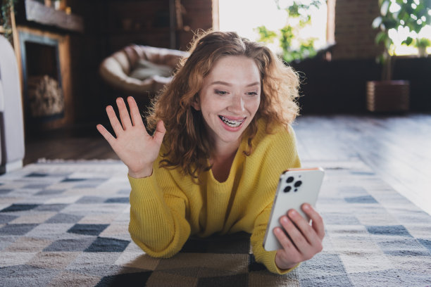 年轻女性躺在地板上的全尺寸照片拿着设备视频通话挥手hi穿着黄色毛衣日光家室内客厅图片下载