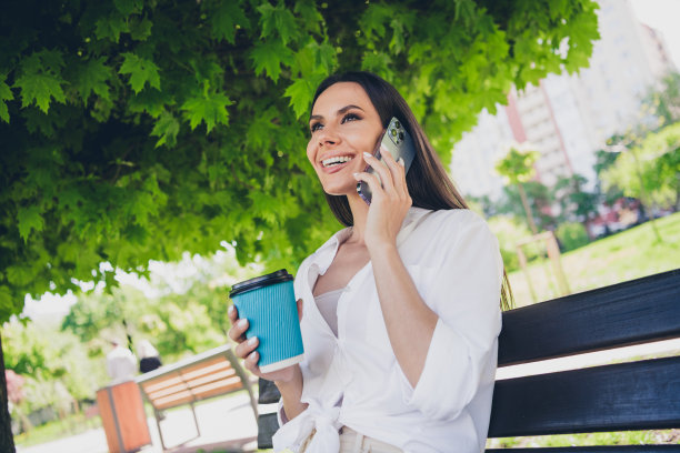 年轻快乐的女人的照片坐在长凳上讲电话喝咖啡穿着白色的衣服散步公园阳光明媚的夏天天气外面图片下载