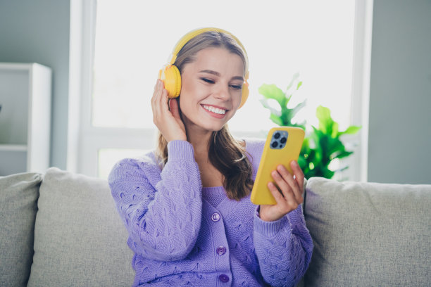 可爱开朗的女孩照片坐在沙发上，耳机拿着小玩意，穿着紫色的衣服在宽敞的房间里消磨时光图片下载