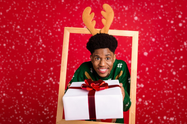 滑稽的家伙的照片出现在框架之外给你礼物包戴鹿头带跳线孤立在红色背景图片下载