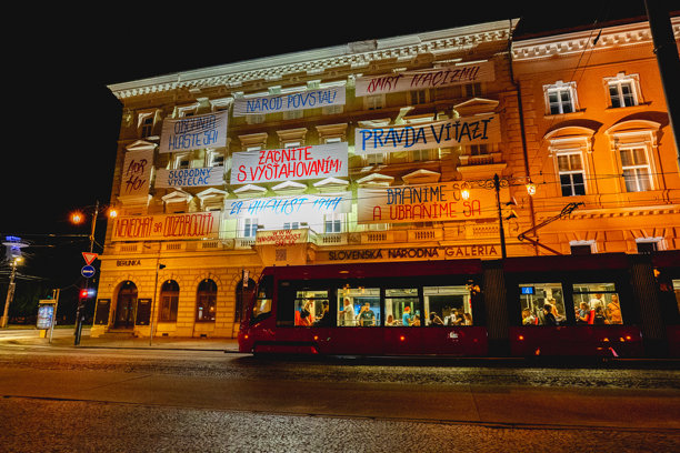 历史悠久的布拉迪斯拉发老城区的夜间照片，有一辆有轨电车和一座展示斯洛伐克民族起义纪念标志的建筑。图片下载