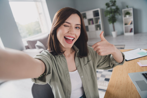 滑稽开朗的人坐在椅子上的照片使自拍眼睛眨眼手臂手指示范拇指在室内工作站图片下载