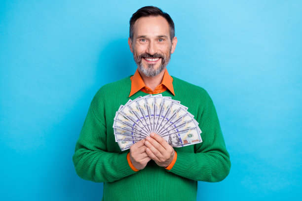 中年微笑的照片穿绿色时髦的套头衫男人深色的头发拿着很多现金钞票美元百万富翁孤立在蓝色的背景图片下载