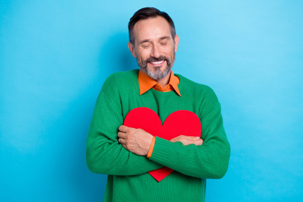 照片正面成熟的老年黑发男子灰胡子微笑满意拥抱红纸情人节形状符号孤立在蓝色背景图片下载