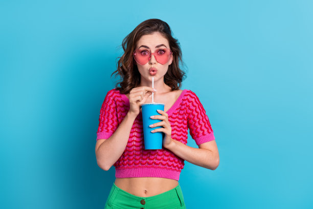 甜美的照片，漂亮可爱的女孩，卷发，穿着粉红色的上衣，撅起的嘴唇，拿着一杯苏打水，孤立在蓝色的背景上图片下载