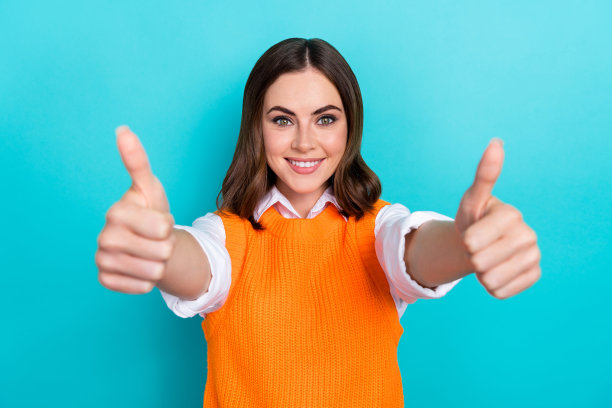 照片中满意的开朗的女人，梳着鲍勃发型，穿着橙色马甲，在蓝绿色的背景上向你竖起大拇指图片下载