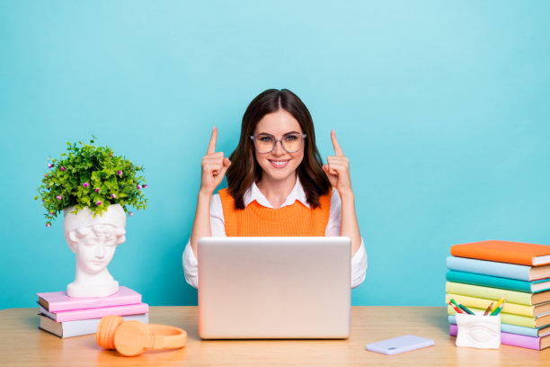 迷人聪明的女士的照片，穿着时髦的衣服，坐在桌子上，许多书，现代设备，在青色背景上孤立的空间图片下载