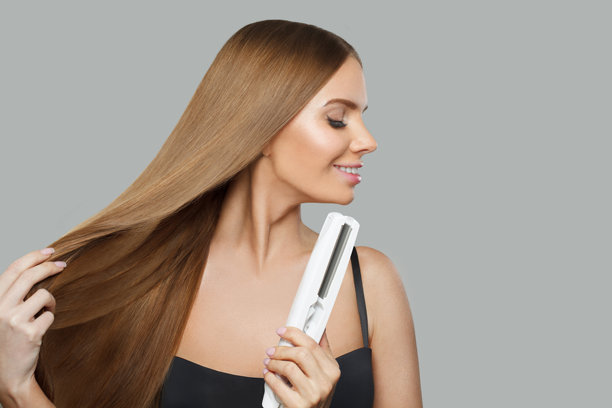 迷人的黑发女人拿着直发棒，在白色的背景上拉直她光滑闪亮的长发。护发、发型、美发及发型设计理念图片下载