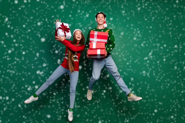 照片中滑稽疯狂的夫妇拿着一堆礼盒享受跳跃飞行戴着帽子丑陋的套头衫孤立的绿色背景图片下载