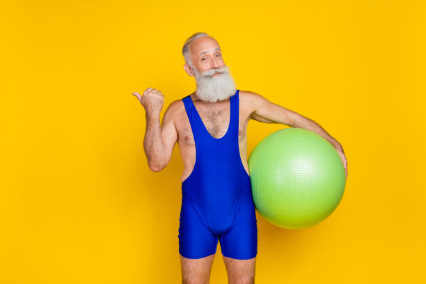 老养老金领取者的肖像照片积极的祖父胡须露出手指空的空间拿着新的健身球兴奋地提供孤立的黄色背景图片下载