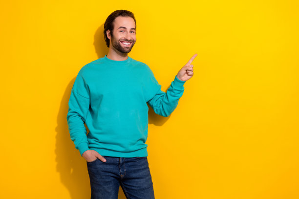 照片的深色头发微笑的男人穿套头衫，把手口袋指导手指模型新的超市促销孤立的黄色背景图片下载