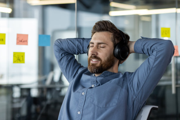 特写照片，年轻的男性上班族戴着耳机坐在工作场所休息，闭着眼睛，双手放在脑后图片下载