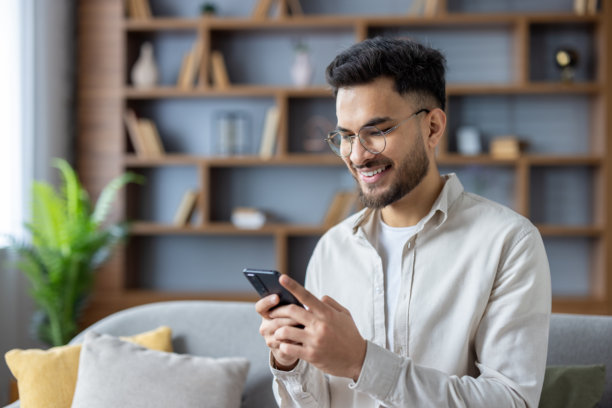 年轻的印度男子坐在家里的沙发上，手里拿着手机微笑的照片图片下载
