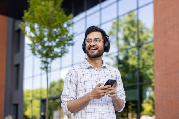 戴眼镜和衬衫的印度年轻男子走在城市街道上，戴着耳机，拿着手机，看向远方特写照片图片下载