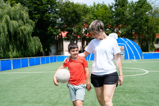 母亲和儿子一起走在足球场上，在晴朗的天空下享受着踢球的时光。图片下载