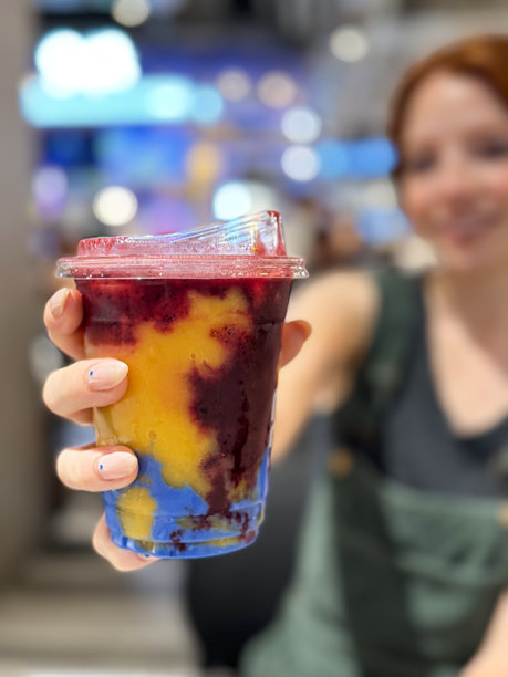 特写照片很吸引人，红头发的女人梳着头发，穿着绿色灯芯绒工作服和黑色露脐上衣，拿着一次性塑料杯，里面装着芒果覆盆子和蓝色螺旋藻奶昔，背景是被照亮的购物中心，重点是前景图片下载