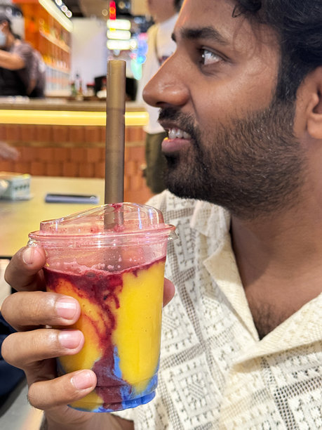 印度男子特写，留有胡须，穿着图案奶油色衬衫，手持塑料一次性盖杯，杯中是芒果覆盆子和蓝色螺旋藻奶昔，配有吸管，背景为明亮的咖啡馆餐厅环境，侧面视角，前景聚焦。图片下载