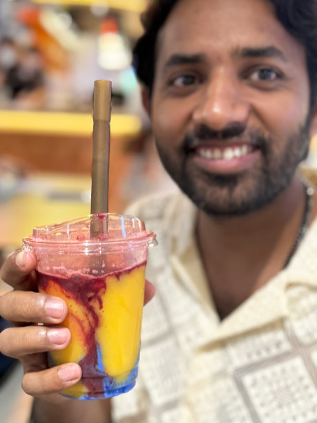 印度男子特写，留有胡须，穿着图案奶油色衬衫，手持塑料一次性盖杯，杯中是芒果覆盆子和蓝色螺旋藻奶昔，插着吸管，正看向镜头，咖啡馆餐厅环境，前景聚焦。图片下载