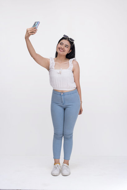 一位漂亮的年轻亚洲女性用手机拍摄了一张向上角度的自拍照。她是一位现代时尚的Z世代女性。全身照片，背景为白色。图片下载