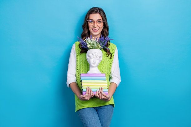 戴眼镜的漂亮年轻女孩手持一堆书籍和雕塑，穿着时尚的绿色衣服，背景为蓝色。图片下载