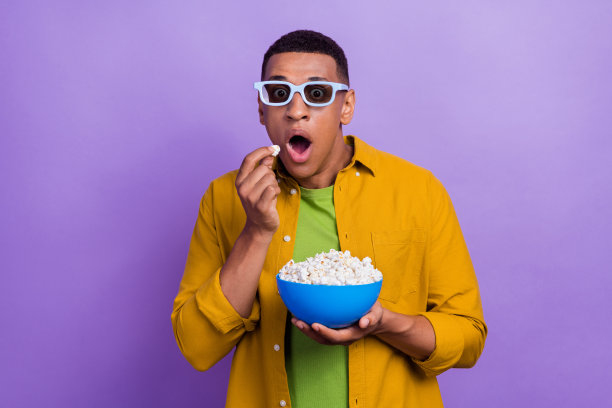 惊讶而印象深刻的帅气男士，穿着黄色衣服，戴着眼镜，兴奋地观看电影，背景为紫色。图片下载