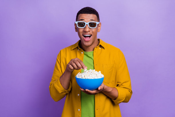 穿着黄色衣服、戴着眼镜的帅气积极的男士在家中观看电影，背景为紫色。图片下载