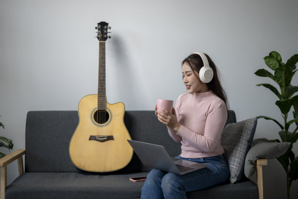 一位女性坐在沙发上，旁边放着一把吉他。她戴着耳机，手里拿着一个杯子。这个场景暗示她正在听音乐或录制歌曲。图片下载