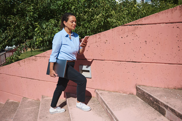 身穿职业装的女性在户外走动，手持笔记本电脑，一边查看智能手机，一边在阳光明媚的日子里攀爬混凝土台阶。图片下载