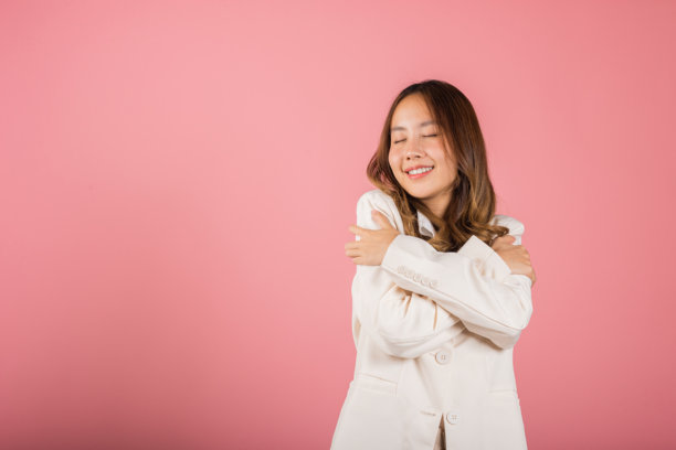 快乐美丽的年轻微笑亚洲女商人抱着自己，回忆瞬间，工作室短片，背景为粉色，关爱自己和自尊的概念图片下载
