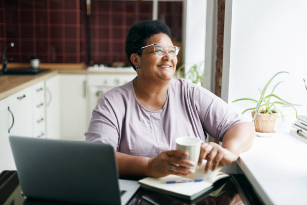 愉快、微笑的超重成熟黑人女性戴着眼镜，坐在厨房桌旁，手中捧着白色咖啡杯，透过窗户欣赏外景，享受热饮，刚刚在笔记本电脑上工作完。图片下载