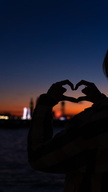 女孩用双手做出心形轮廓，背景是模糊的城市和海洋，黄昏时分的日落，留白空间。竖版照片图片下载