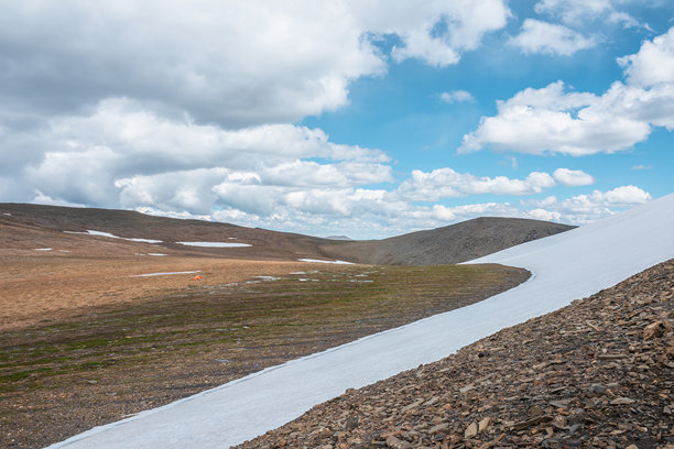 阳光明媚的阿尔卑斯山谷中，一顶鲜艳的橙色帐篷矗立在雪白的冰川旁，云朵在蓝天中飘动。风景如画的高山石径上，孤独的帐篷显得格外醒目。多变的天气在岩石山脉中交替。图片下载