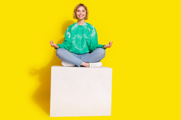 穿着绿色卫衣的快乐漂亮女士在空旷的黄色背景下坐在平台上练习瑜伽的全身照片图片下载