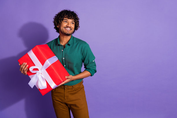 有趣的墨西哥男士的照片肖像，留着波浪形发型，穿着休闲衬衫，在紫色背景下准备生日礼物盒。图片下载