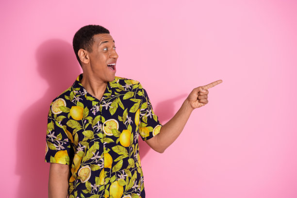 年轻帅气男士的照片肖像，穿着时尚的热带印花服装，指向空旷的空间，背景为粉色。图片下载