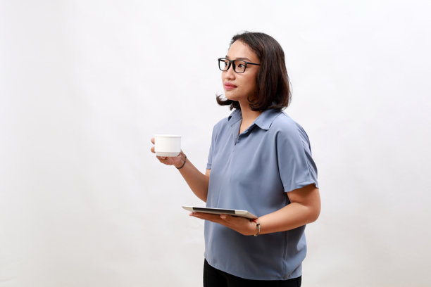 迷人的女商人站在白色背景前，喝着咖啡，使用数字平板，目光投向空旷的空间。图片下载