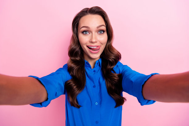 一位满意、心情愉快的女性，卷发，穿着蓝色衬衫，自拍时露出牙齿，背景为粉色。图片下载