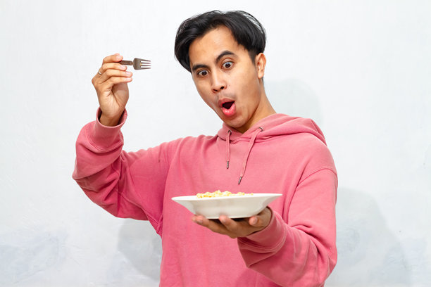 年轻的亚洲男子穿着粉色连帽衫，面带快乐和兴奋的表情，凝视着一盘方便炒面，背景为白色。图片下载