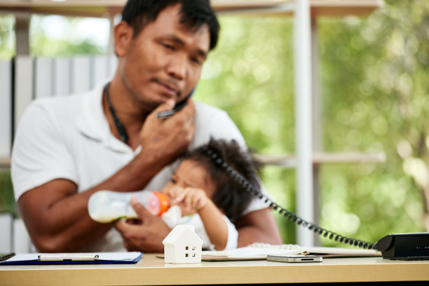 亚洲父亲在桌子上喂宝宝并打电话，平衡工作与育儿。图片下载