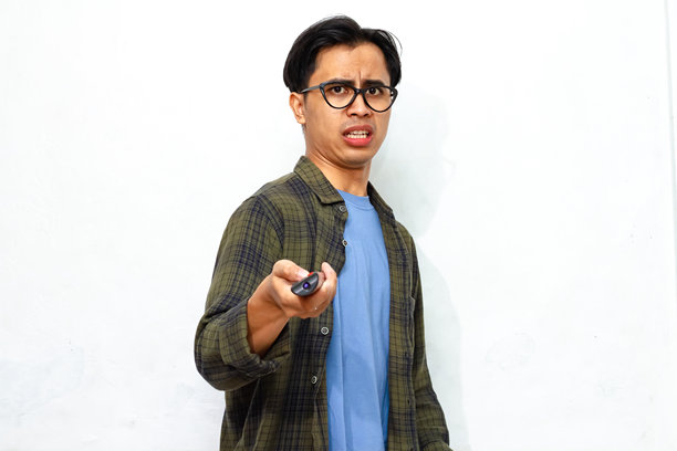 年轻的亚洲男子穿着休闲服，手持遥控器，露出搞笑的震惊表情，背景为白色。图片下载