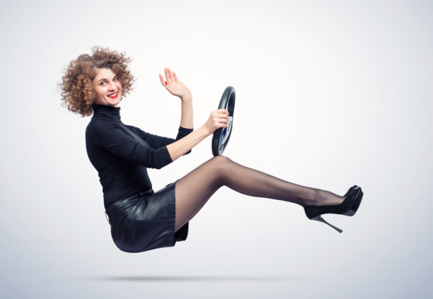 年轻微笑的女性穿着黑色高领毛衣和裙子，一只手握着方向盘，另一只手在打招呼，背景为浅色。图片下载